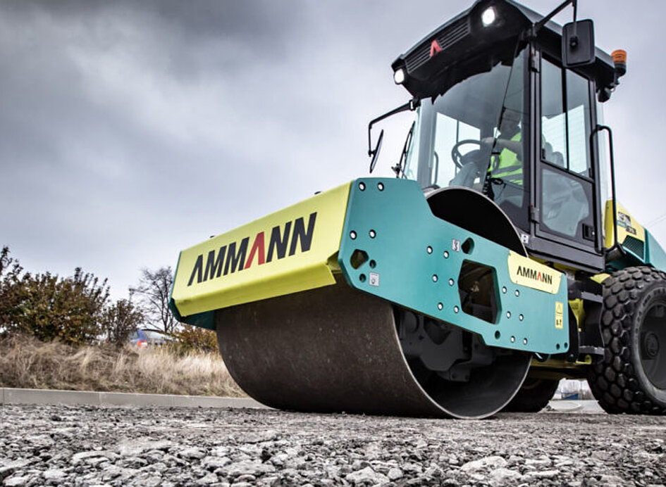 Straßenwalze von Ammann auf einer Baustelle im Einsatz. Die grün-gelbe Maschine verdichtet frischen Asphalt, im Hintergrund sind Bäume und ein grauer Himmel zu sehen.
