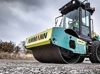 Straßenwalze von Ammann auf einer Baustelle im Einsatz. Die grün-gelbe Maschine verdichtet frischen Asphalt, im Hintergrund sind Bäume und ein grauer Himmel zu sehen.