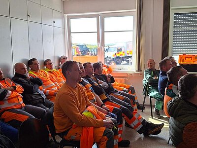 Mehrere Personen sitzen in einem Schulungsraum und hören einem Vortrag zu. Viele tragen orangefarbene Arbeits- und Warnkleidung