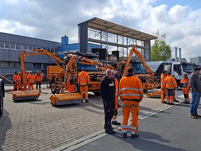 Auf einem Betriebshof stehen mehrere Kommunalfahrzeuge mit Mäh- und Reinigungsanbauten. Viele Personen in orangefarbener Warnkleidung tauschen sich zwischen den Fahrzeugen aus