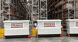 Routenzug: Linde Elektroschlepper zieht beladene STRABAG-Transportwagen durch ein Hochregallager.