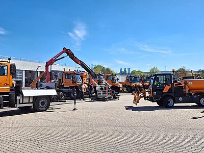 Mehrere orangefarbene Kommunalfahrzeuge mit Arbeitsgeräten stehen auf einem Betriebshof. Einige Personen in Warnkleidung besichtigen die ausgestellten Fahrzeuge