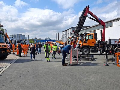 Personen in Warnkleidung stehen auf einem Betriebsgelände zwischen ausgestellten Kommunalfahrzeugen. Im Vordergrund wird ein Anbaugerät an einem Fahrzeug betrachtet