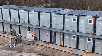 Zweistöckige Containeranlage der Firma Beutlhauser mit umlaufendem Laufsteg und Außentreppe auf einer Baustelle, davor Baumaterial und Werkzeugkisten auf einem Holzlaufweg.
