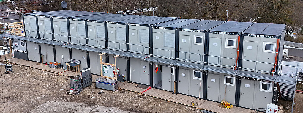 Zweistöckige Containeranlage der Firma Beutlhauser mit umlaufendem Laufsteg und Außentreppe auf einer Baustelle, davor Baumaterial und Werkzeugkisten auf einem Holzlaufweg.