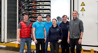 Eine Gruppe von Mitarbeitenden steht lächelnd vor einem geöffneten technischen Großgerät mit sichtbaren Kabel- und Batteriesystemen in einer Werkhalle.