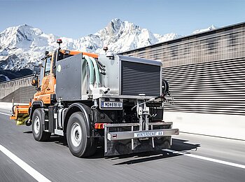 Orangefarbenes Winterdienstfahrzeug mit Sole-Sprühanlage von Ecotech auf Autobahn, im Hintergrund verschneite Berge