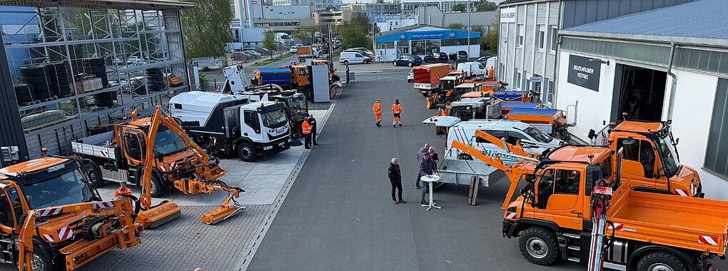 Blick von oben auf einen Betriebshof mit mehreren orangefarbenen Kommunalfahrzeugen und Arbeitsgeräten. Einzelne Personen stehen zwischen den ausgestellten Fahrzeugen