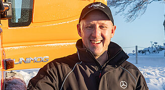Ein Man neben einem organgenfarbenen Unimog und Schnee im Hintergrund.