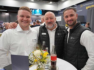 Drei Menschen blicken in die Kamera, im Hintergrund ein Messestand.