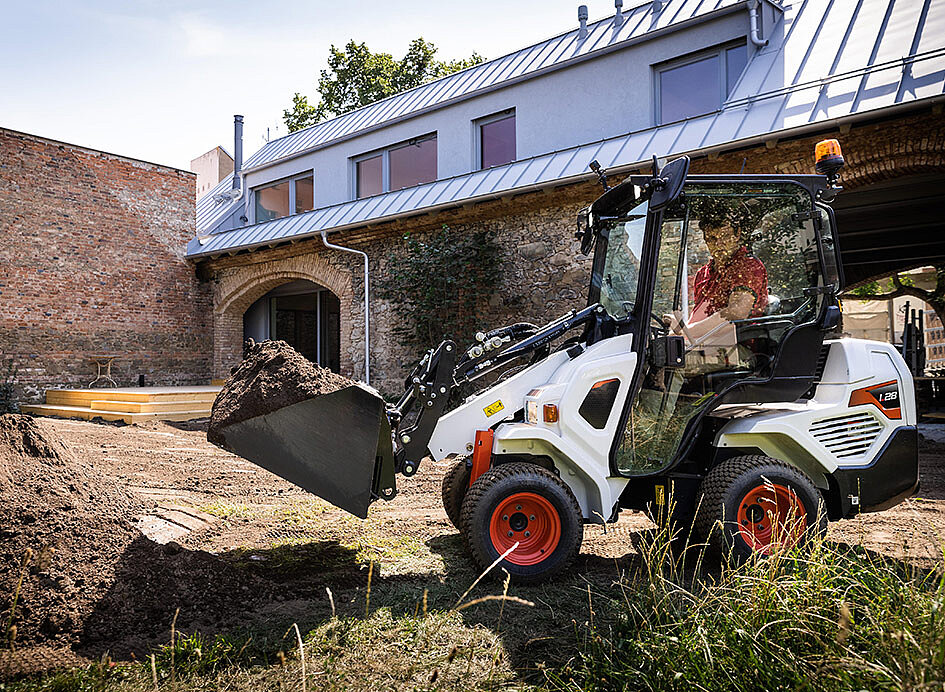 Bobcat Kompakt-Knicklader befördert eine Schaufel Erde vor einem Haus