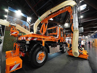 Ein orangefarbener Unimog mit Anbaugeräten auf einem Messestand in einer Halle.