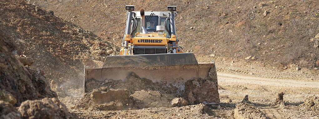 Eine gelbe Liebherr-Planierraupe schiebt auf einer Erdbaustelle mit ihrem breiten Schild lockeres Erdreich vor sich her.