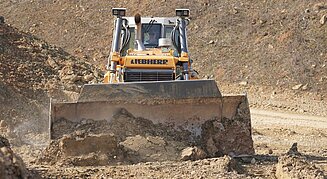 Eine gelbe Liebherr-Planierraupe schiebt auf einer Erdbaustelle mit ihrem breiten Schild lockeres Erdreich vor sich her.