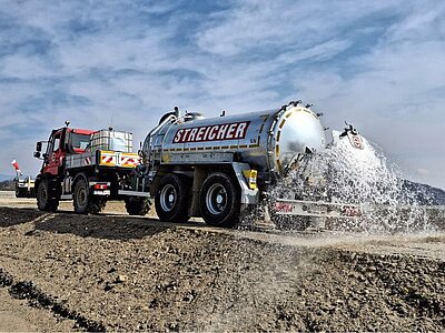 Rotes Kommunalfahrzeug mit silbernem Tankanhänger verteilt Wasser auf einer unbefestigten Fläche
