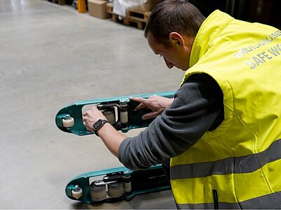 Ein Mitarbeiter in Warnweste prüft die Rollen eines Hubwagens in einer Lagerhalle.
