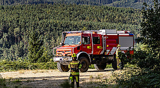 Ein rot-weißer Unimog im bewaldeten Gelände mit zwei Feuerwehrleuten mit Drohne vor dem Fahrzeug.
