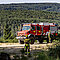 Ein rot-weißer Unimog im bewaldeten Gelände mit zwei Feuerwehrleuten mit Drohne vor dem Fahrzeug.