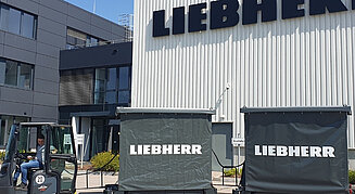 Außenansicht eines modernen Industriegebäudes mit großem „LIEBHERR“-Schriftzug; im Vordergrund ein Gabelstapler mit zwei angehängten Planenwagen, Fahrer im Fahrzeug