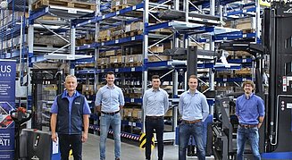 Sechs Männer stehen vor Hochregallagern mit blauen Metallträgern in einer Lagerhalle; links ein Roll-up mit dem Logo „QUADRUS Metalltechnik“ und Informationen zum Leistungsspektrum