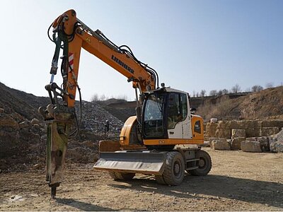 Liebherr-Mobilbagger mit Hydraulikhammer in einem Steinbruch, im Hintergrund gesägte Steinblöcke.