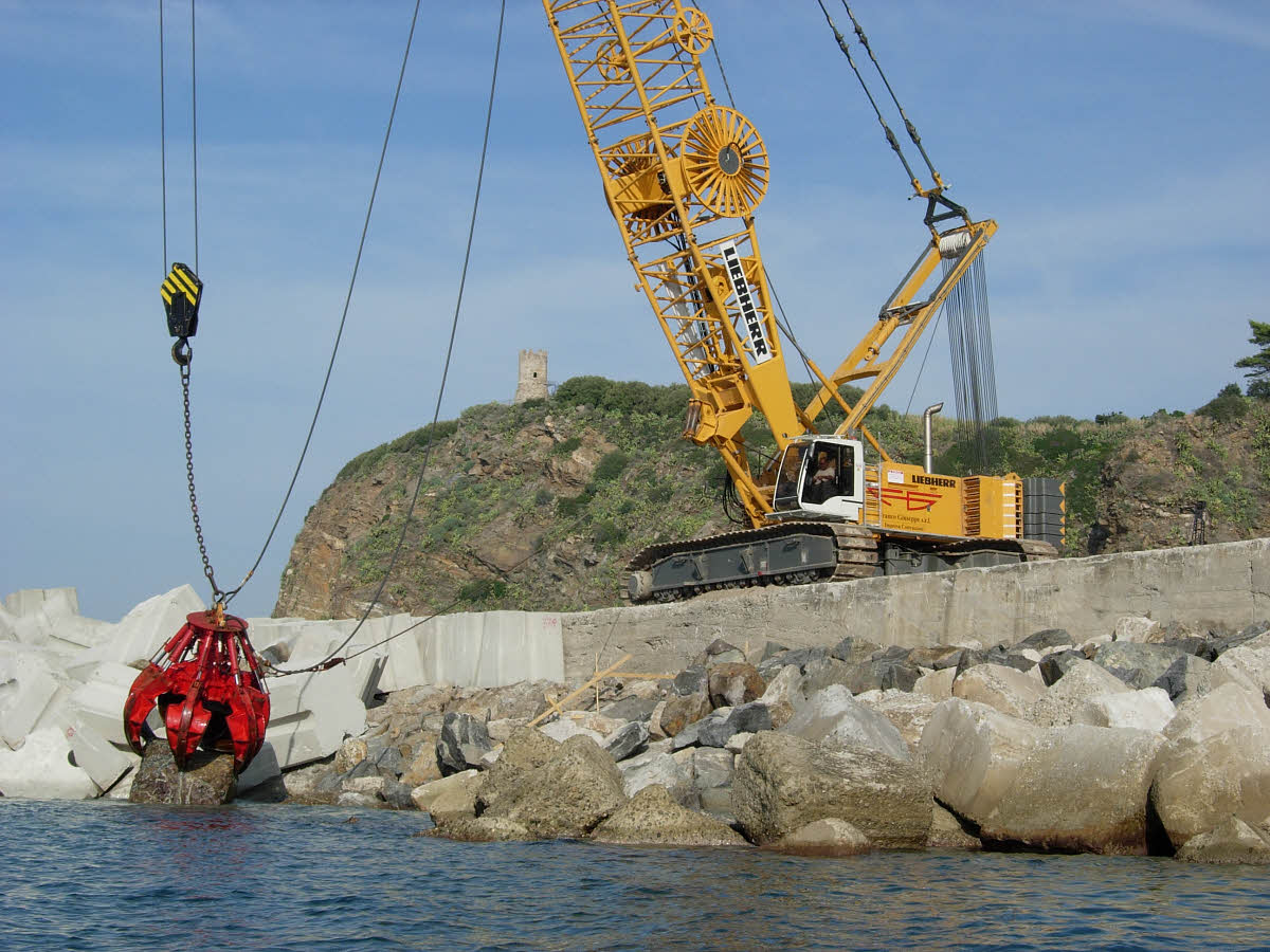Ein Liebherr Seilbagger hebt mit Hilfe einer Greifzange einen großen Stein aus dem Meer. 