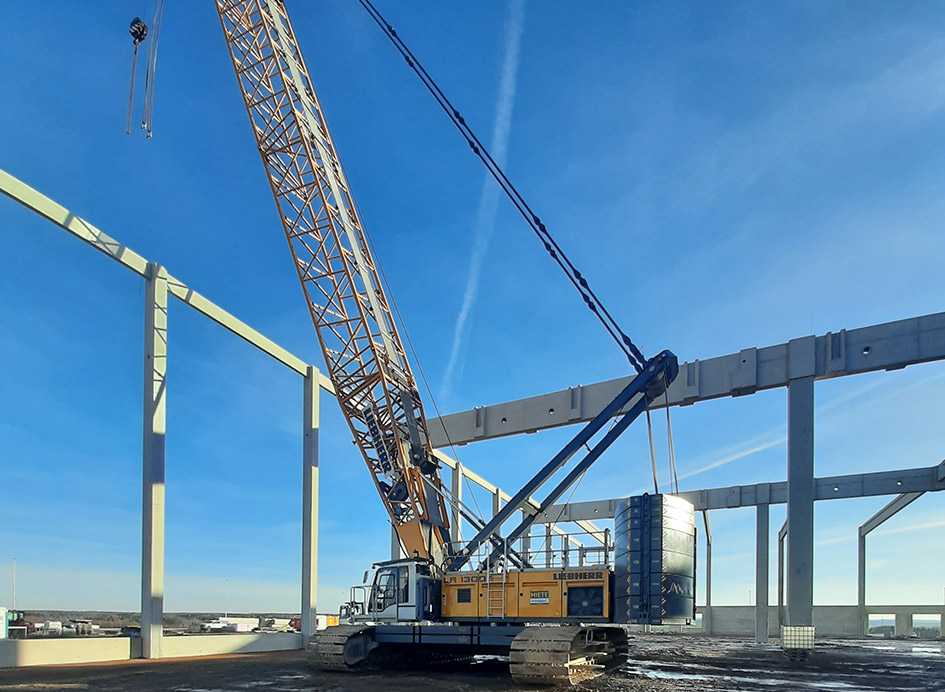 Ein gelber Raupenkran auf einer Baustelle einer Halle unter blauem Himmel.