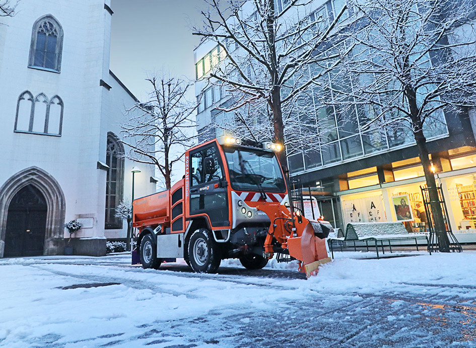 Ein orangenfarbenes Reform Fahrzeug mit Anbaugeräten auf einem verschneiten Stadtplatz.