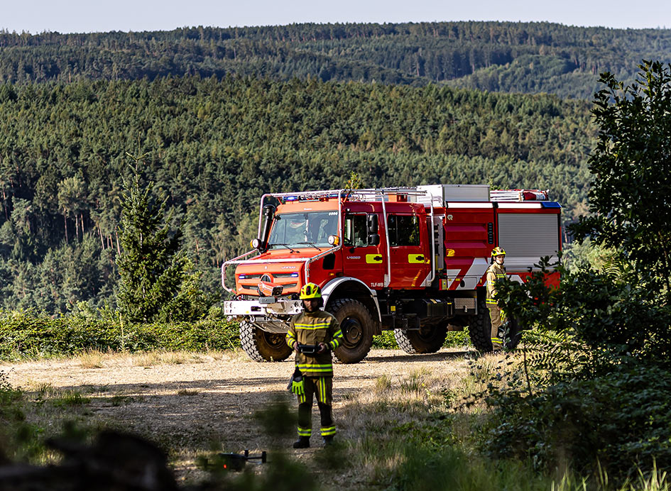 Ein rot-weißer Unimog im bewaldeten Gelände mit zwei Feuerwehrleuten mit Drohne vor dem Fahrzeug.