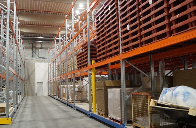 Lagerhalle mit Hochregalen aus Metall, gefüllt mit roten Kunststoffpaletten und verschiedenen Kartonagen, Blick in einen langen Gang mit Betonboden